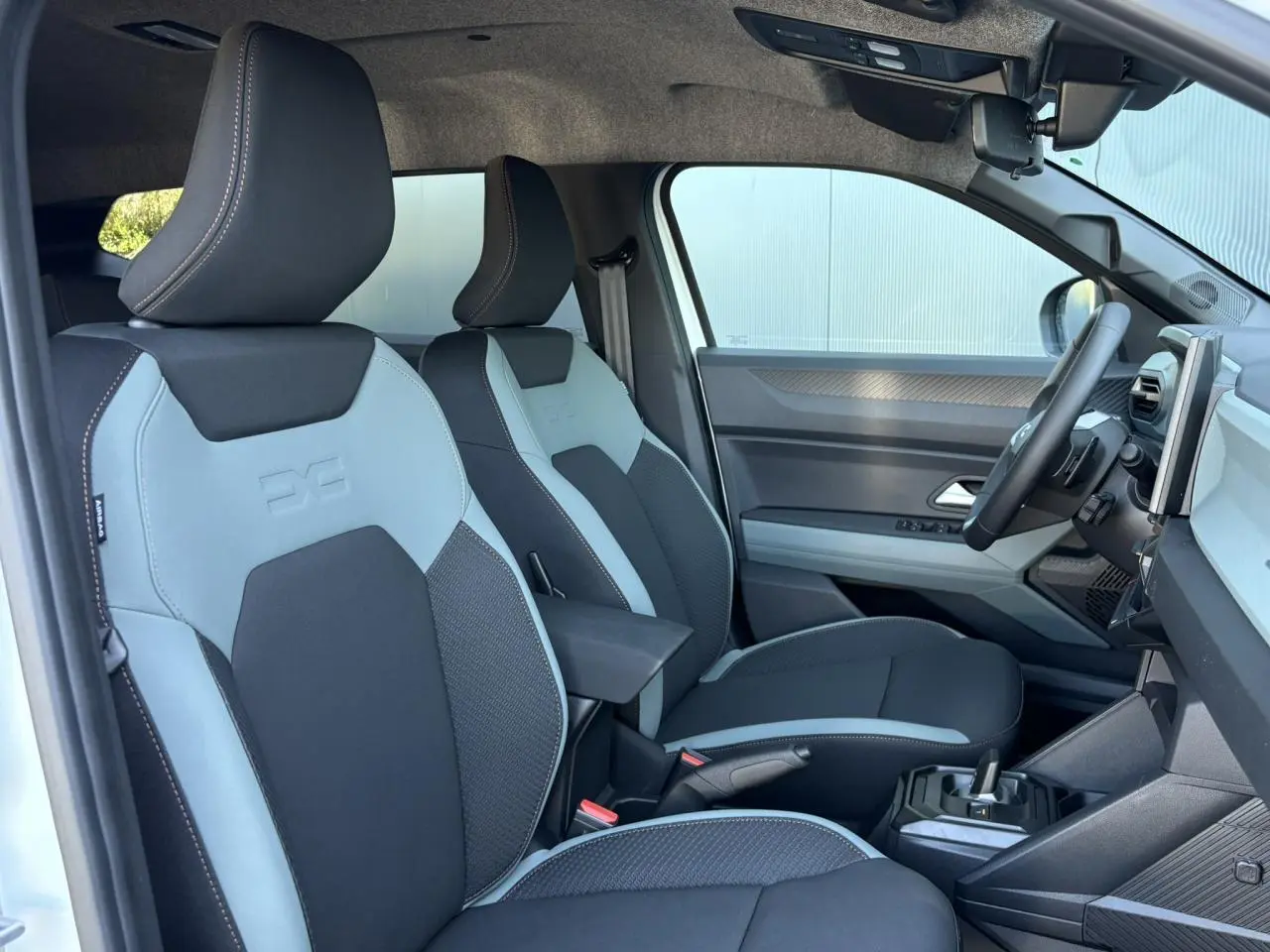 Vue intérieure côté conducteur du Dacia Duster 2026, sièges bi-ton gris clair et noir avec accoudoir central.