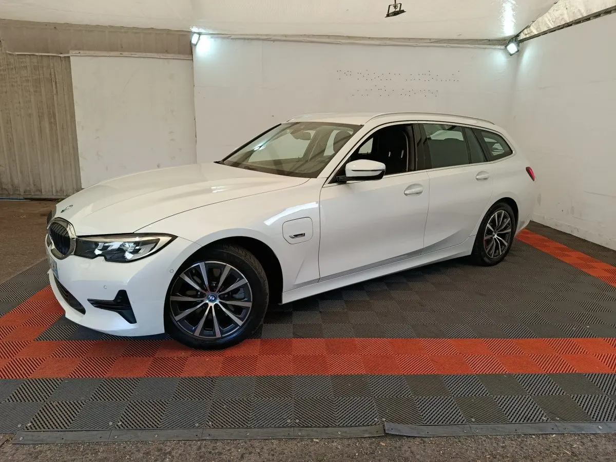 BMW Série 3 320e blanche vue 3/4 avant droit en break, avec jantes alliage et prise de recharge latérale visible.