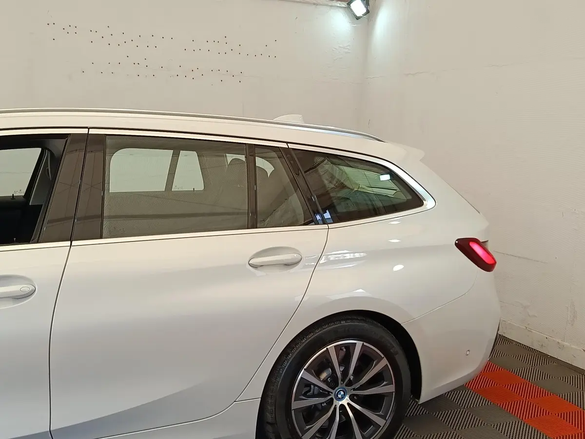 Vue latérale droite d'une BMW Série 3 Touring hybride blanche avec feux arrière allumés dans un garage.