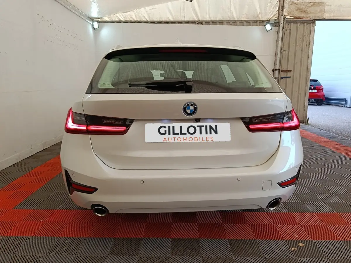 Vue arrière d'une BMW Série 3 320e blanche avec feux LED allumés, double sortie d'échappement et logo BMW visible.
