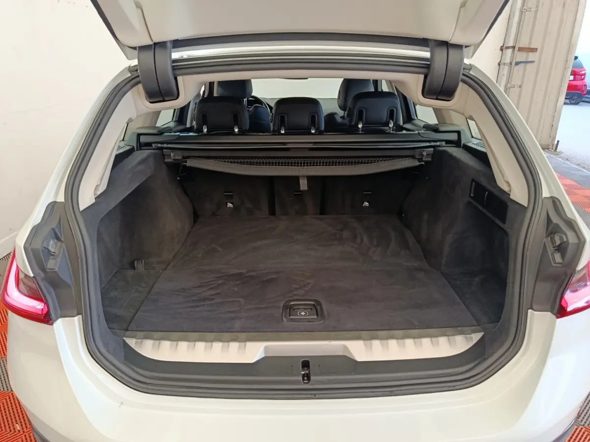 Coffre ouvert vue arrière d’une BMW Série 3 320e blanche, espace de chargement avec tapis noir et banquette rabattable.