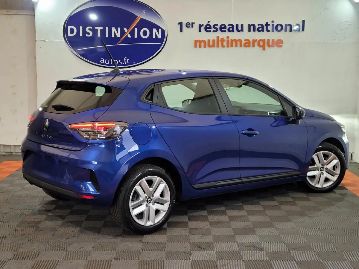 Vue 3/4 arrière droite d'une Renault Clio bleu 2025 stationnée en intérieur avec feux arrière allumés.