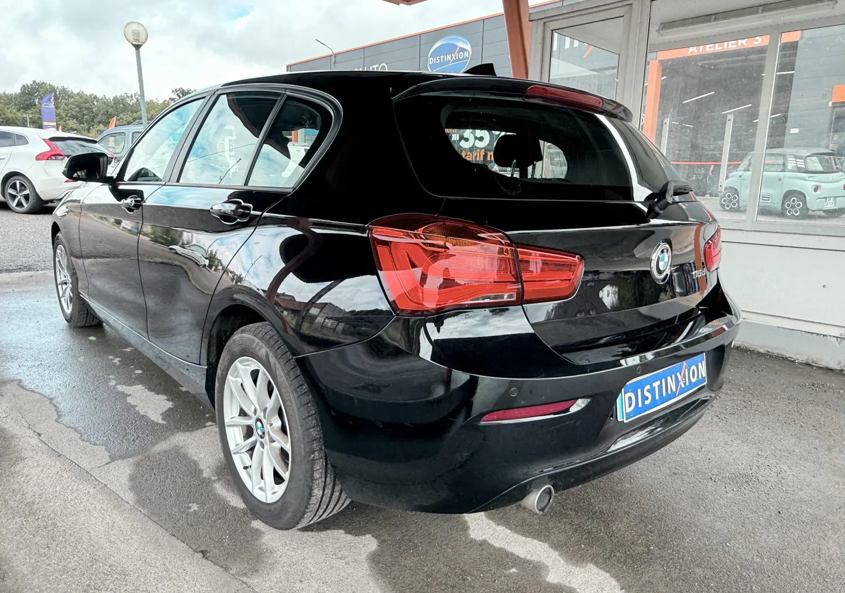 BMW Série 1 noire vue en 3/4 arrière droit, avec feux arrière rouges et jantes argentées, stationnée sous abri.
