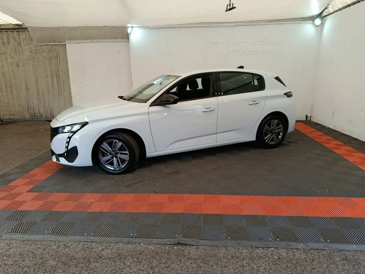Peugeot 308 blanche vue de profil côté gauche, dans un espace fermé avec sol gris et bandes rouges.