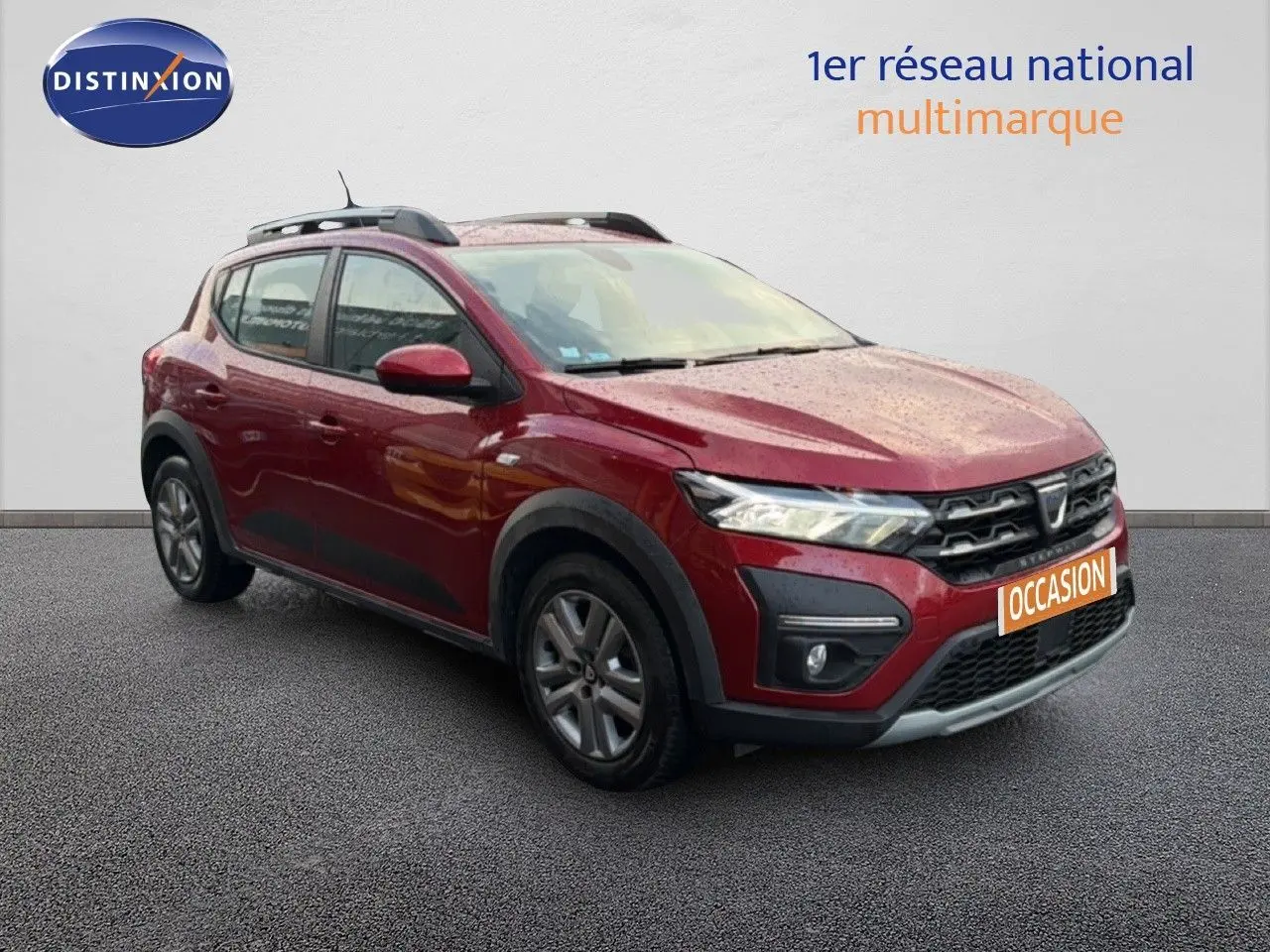 Dacia Sandero Stepway rouge fusion vue 3/4 avant gauche, avec barres de toit et protections noires.
