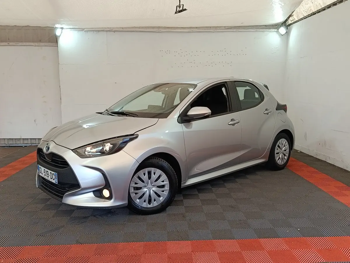Toyota Yaris hybride gris clair vue 3/4 avant droit dans un garage avec feux allumés et jantes standard.