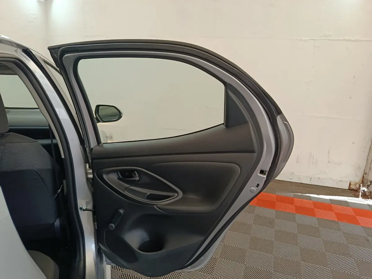 Vue intérieure de la porte arrière droite ouverte d'une Toyota Yaris hybride gris clair, montrant la garniture noire et la manivelle de vitre.