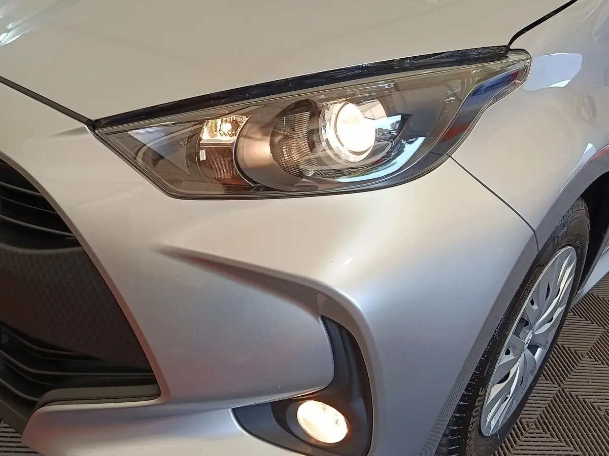 Gros plan sur l'avant droit gris clair de la Toyota Yaris hybride 2022, mettant en valeur les phares allumés et le feu antibrouillard.