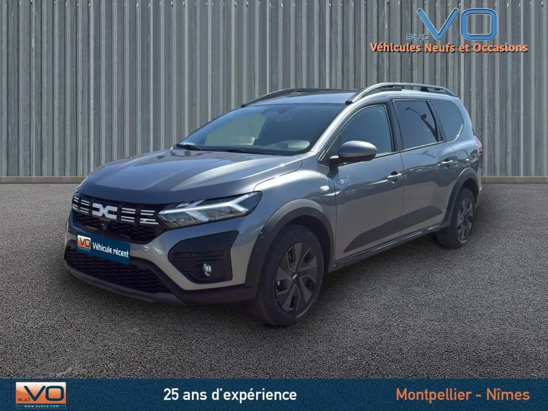Dacia Jogger gris en vue 3/4 avant droit, avec barres de toit et jantes noires, stationné devant un mur métallique.