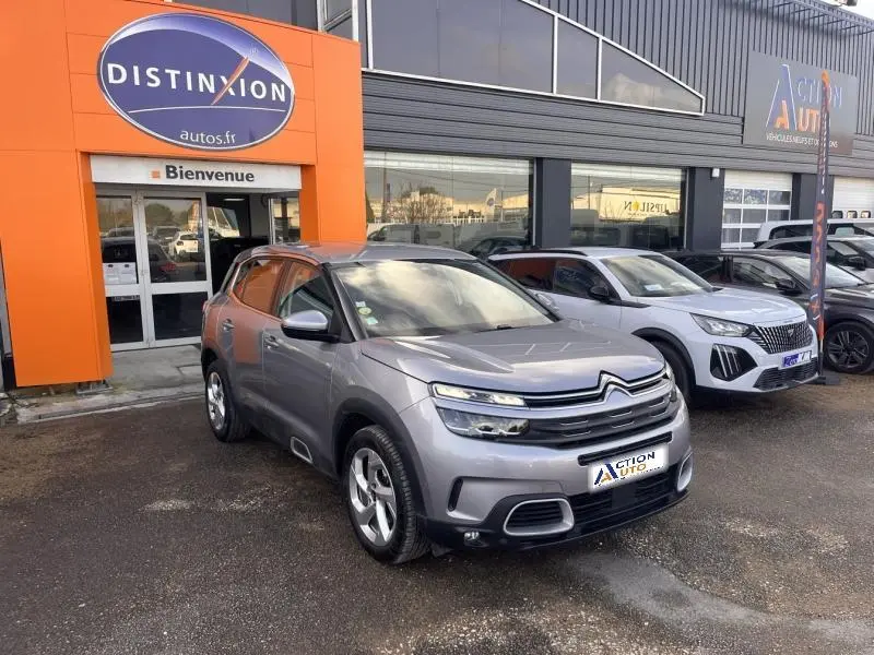 Vue 3/4 avant d'un Citroën C5 Aircross gris Platinium stationné devant un garage avec jantes alliage et feux LED.