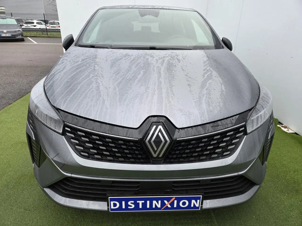 Vue avant d'une Renault Clio V gris schiste métal avec calandre noire et logo Renault proéminent, carrosserie mouillée.