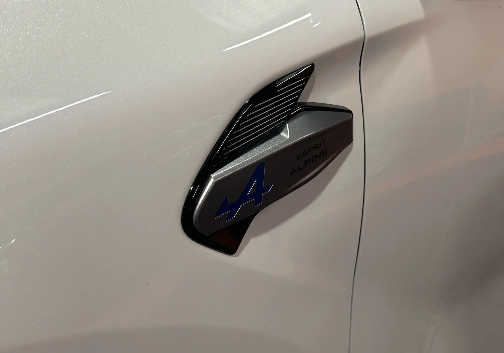 Gros plan sur l'écusson Esprit Alpine bleu et noir sur la porte blanche nacrée d'un Renault Captur II Ph.2.