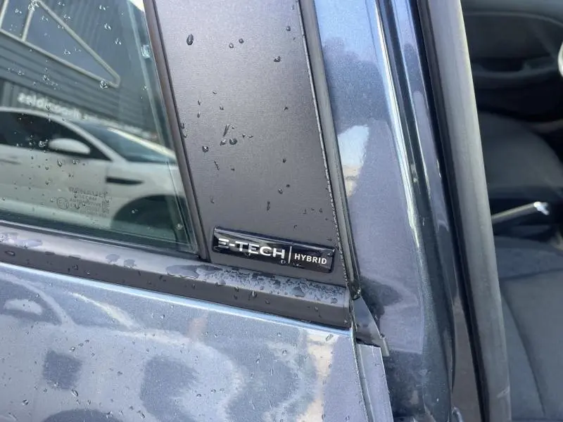 Gros plan sur l'emblème E-Tech Hybrid sur la porte côté gauche d'une Renault Clio gris Titanium avec gouttes d'eau.