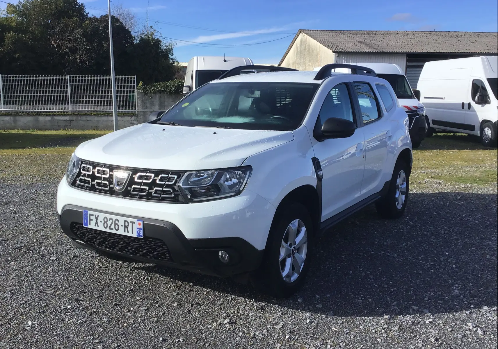 Dacia Duster blanc en 3/4 avant droit, avec calandre noire et jantes alliage 16 pouces, stationné sur un sol gravillonné.