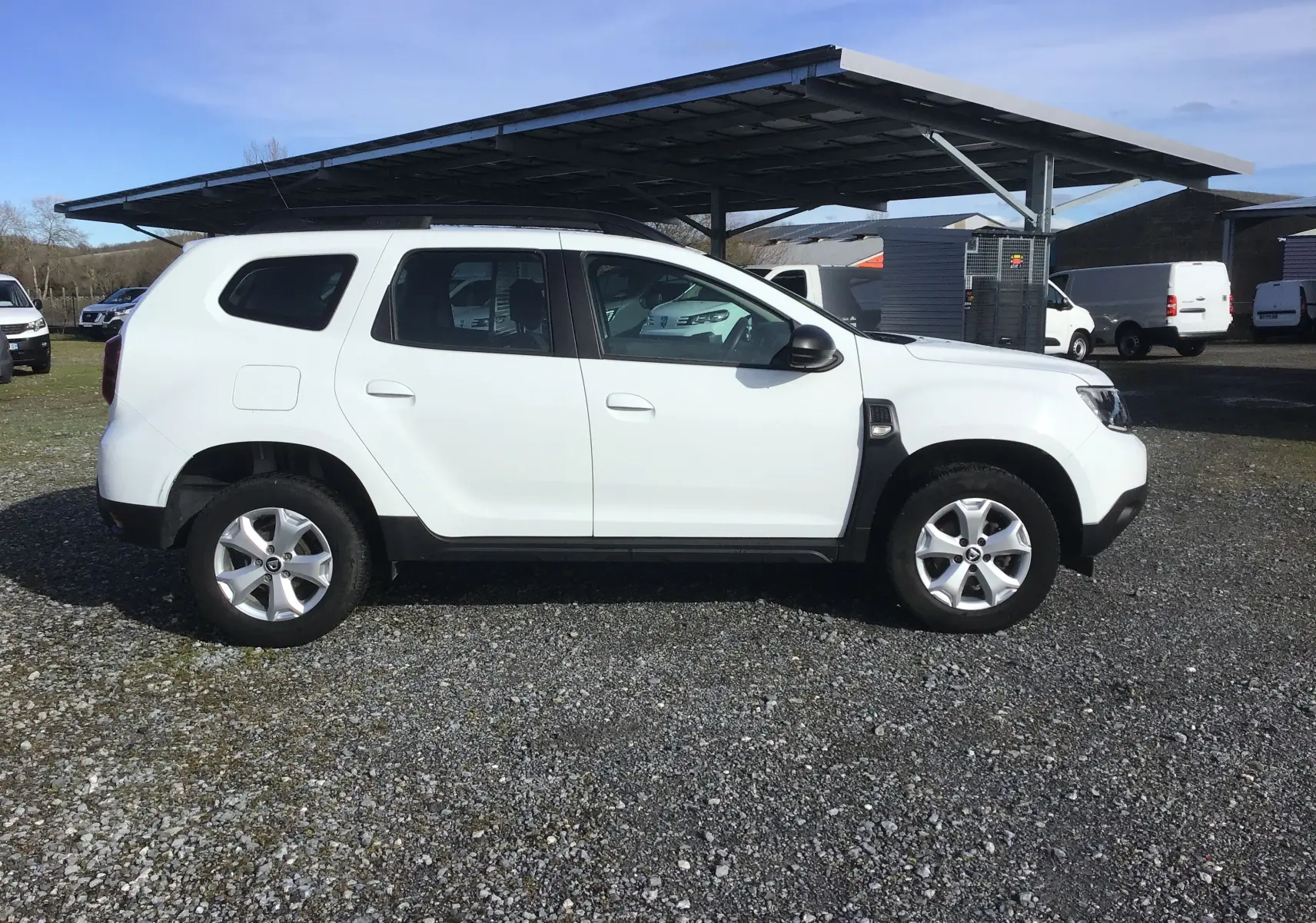 Profil côté gauche d'un Dacia Duster blanc 2021, version 4x2, avec jantes alliage et toit noir sous un abri extérieur.