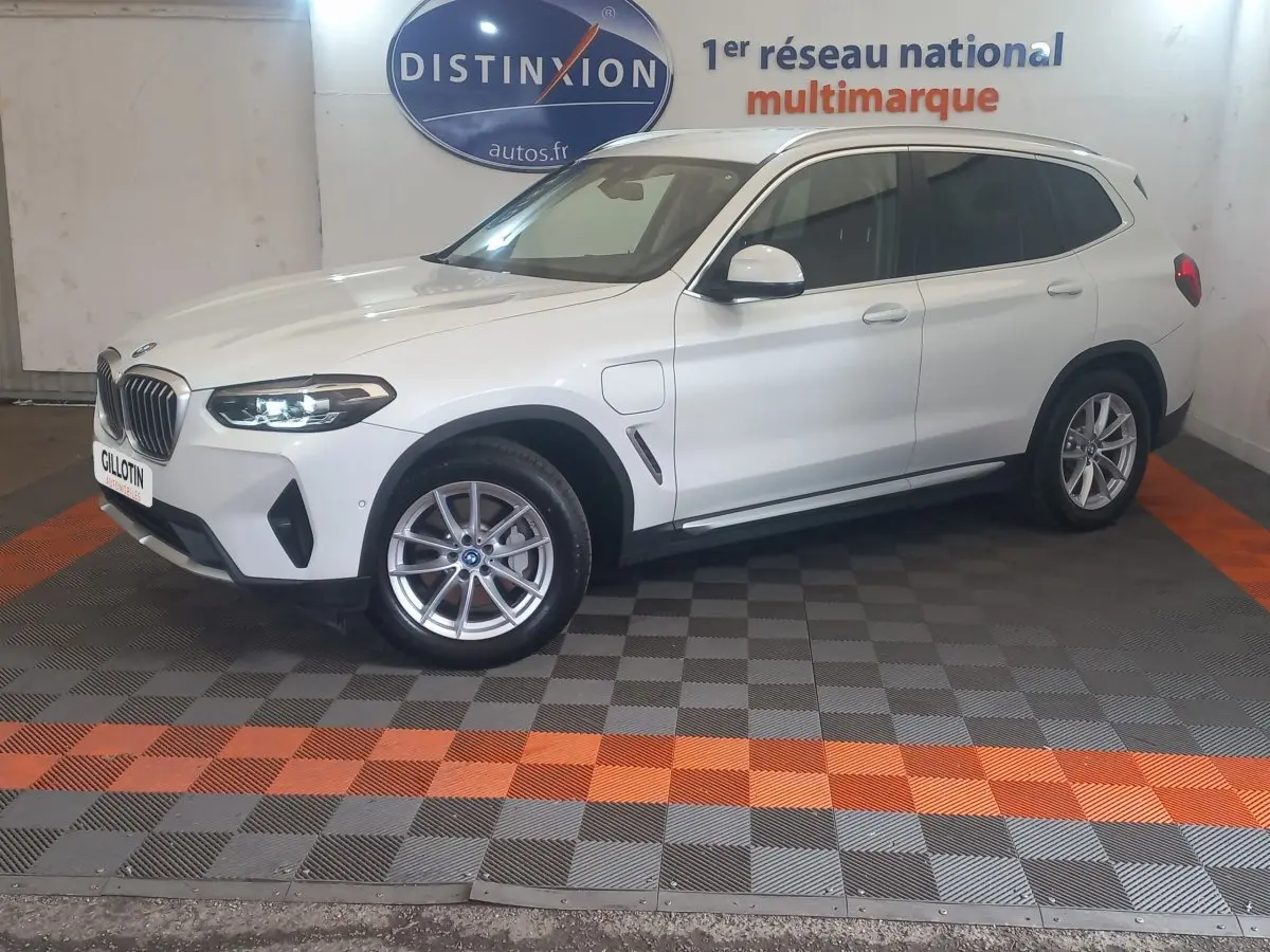 BMW X3 xDrive30e blanc en vue 3/4 avant droit, avec phares LED allumés et jantes alliage visibles.