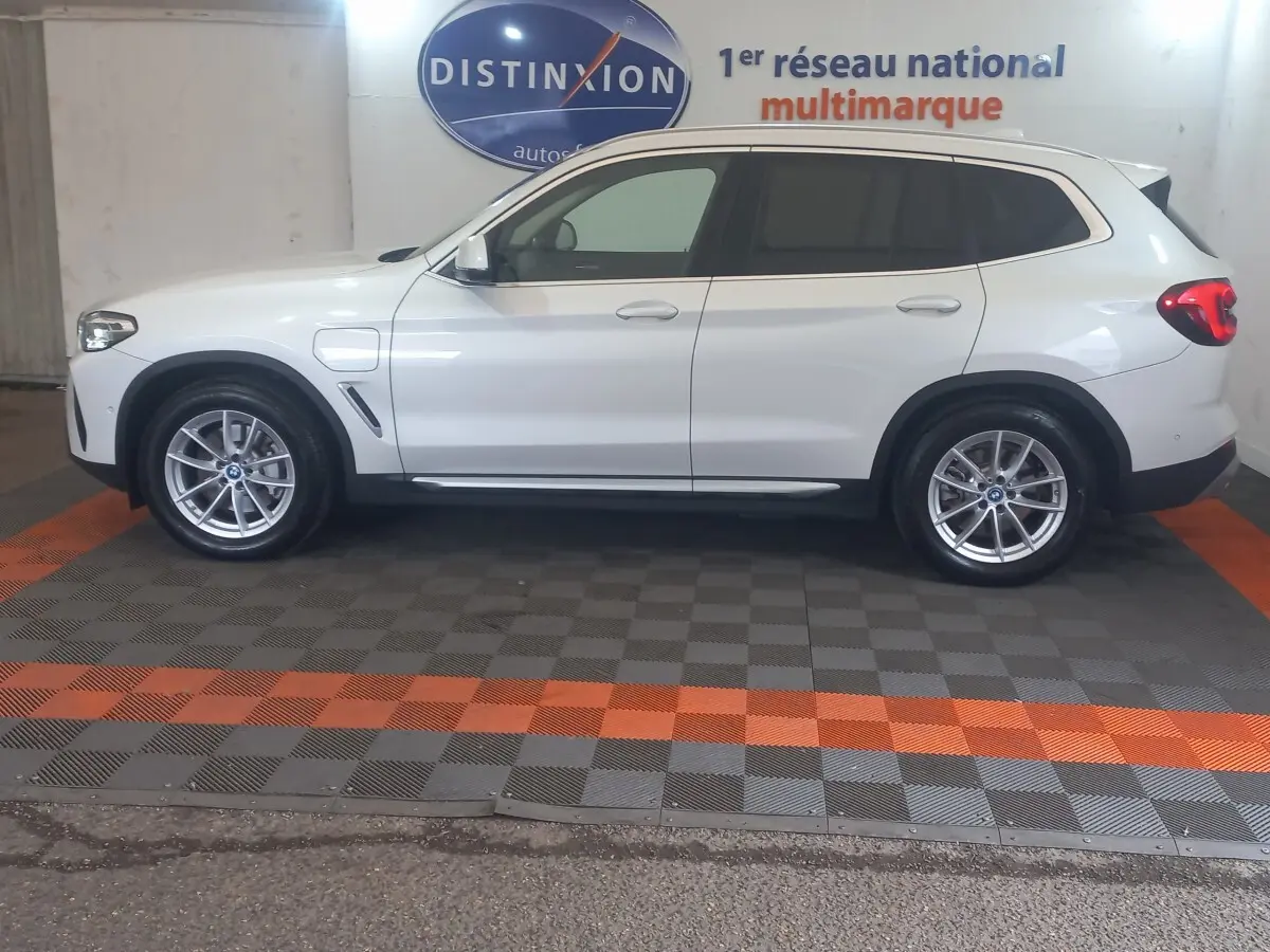 Vue de profil côté gauche d'un BMW X3 xDrive30e blanc, hybride rechargeable, avec jantes alliage et prises de charge visibles.