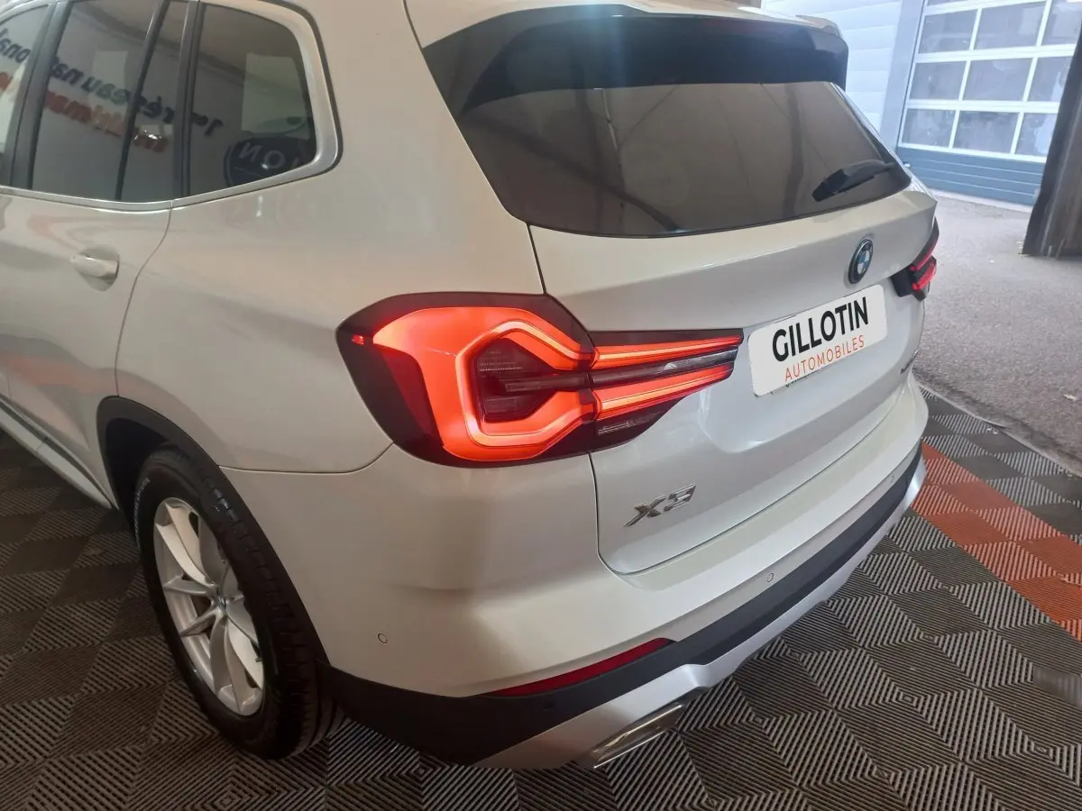 Vue 3/4 arrière gauche d’un BMW X3 blanc avec feux arrière allumés et logo visible sur coffre.