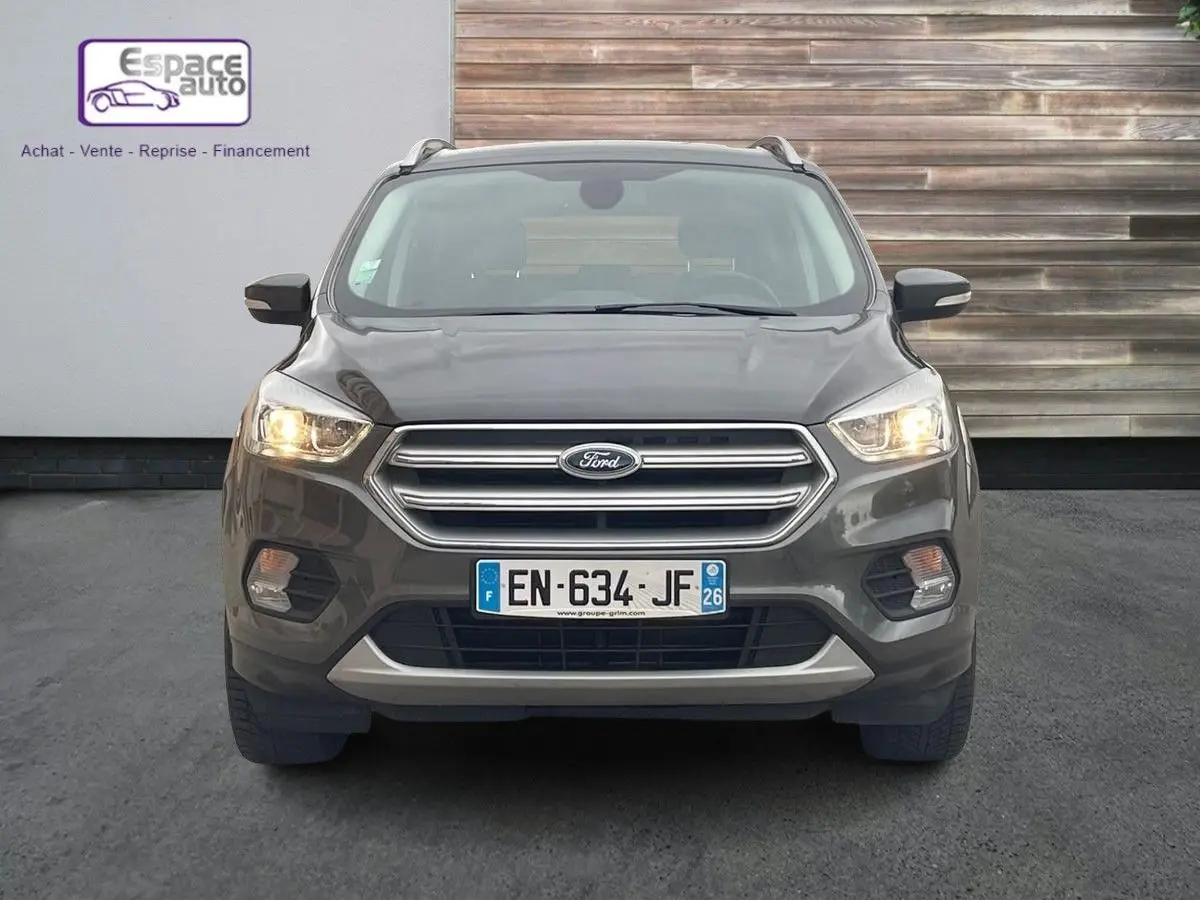 Vue frontale d'un Ford Kuga gris 2017 avec phares allumés et calandre chromée, stationné devant un mur en bois.