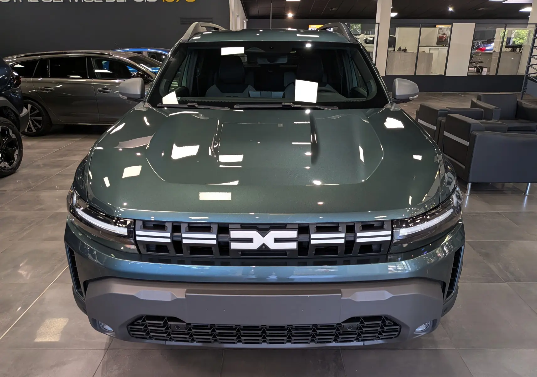 Vue frontale d'un Dacia Duster Hybrid-G 150 CH 4x4 en Vert Cèdre, avec calandre noire et détails modernes en showroom.