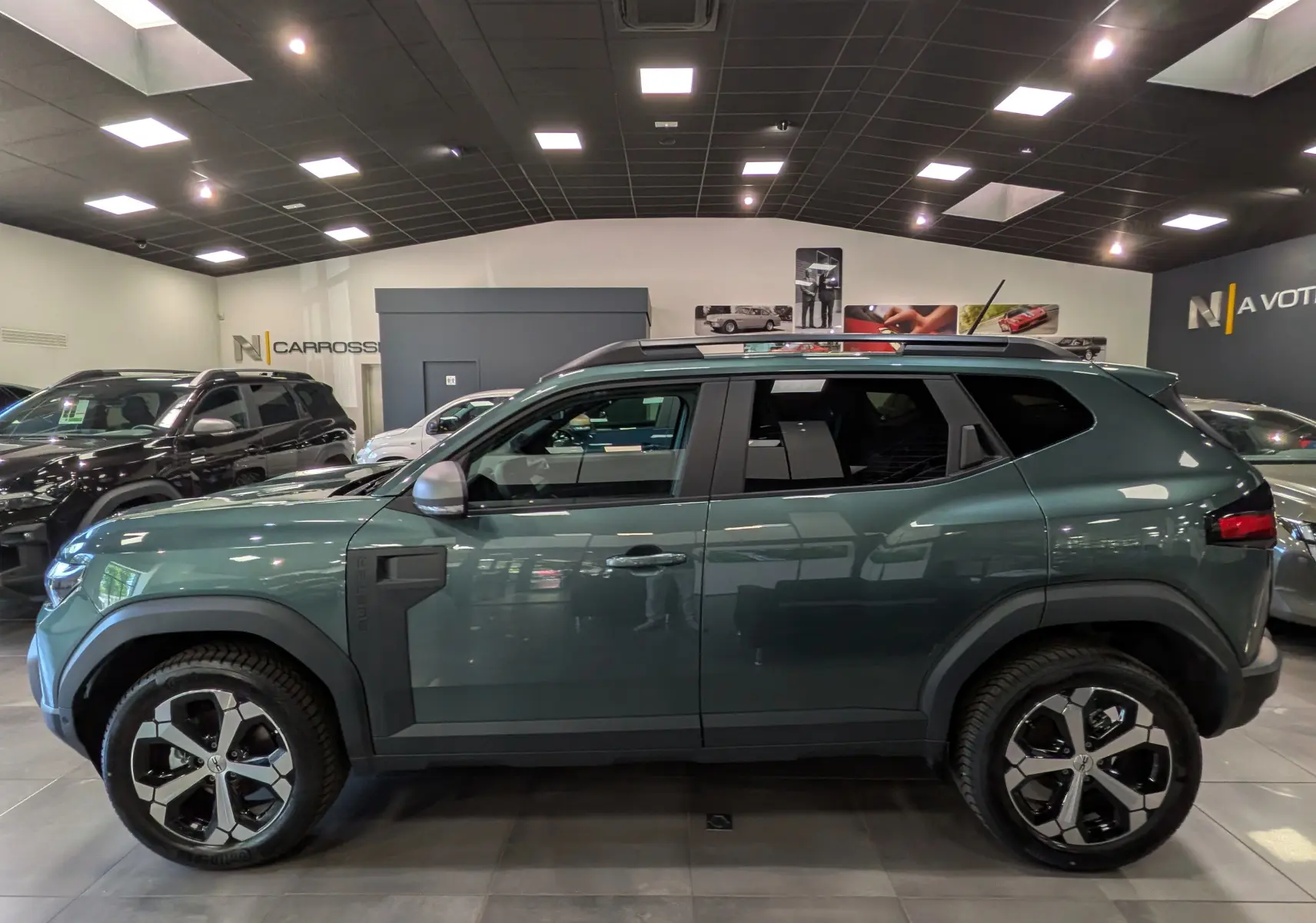 Vue de profil côté gauche du Dacia Duster Hybrid-G 150 CH 4x4 en Vert Cèdre avec jantes bi-ton et barres de toit.