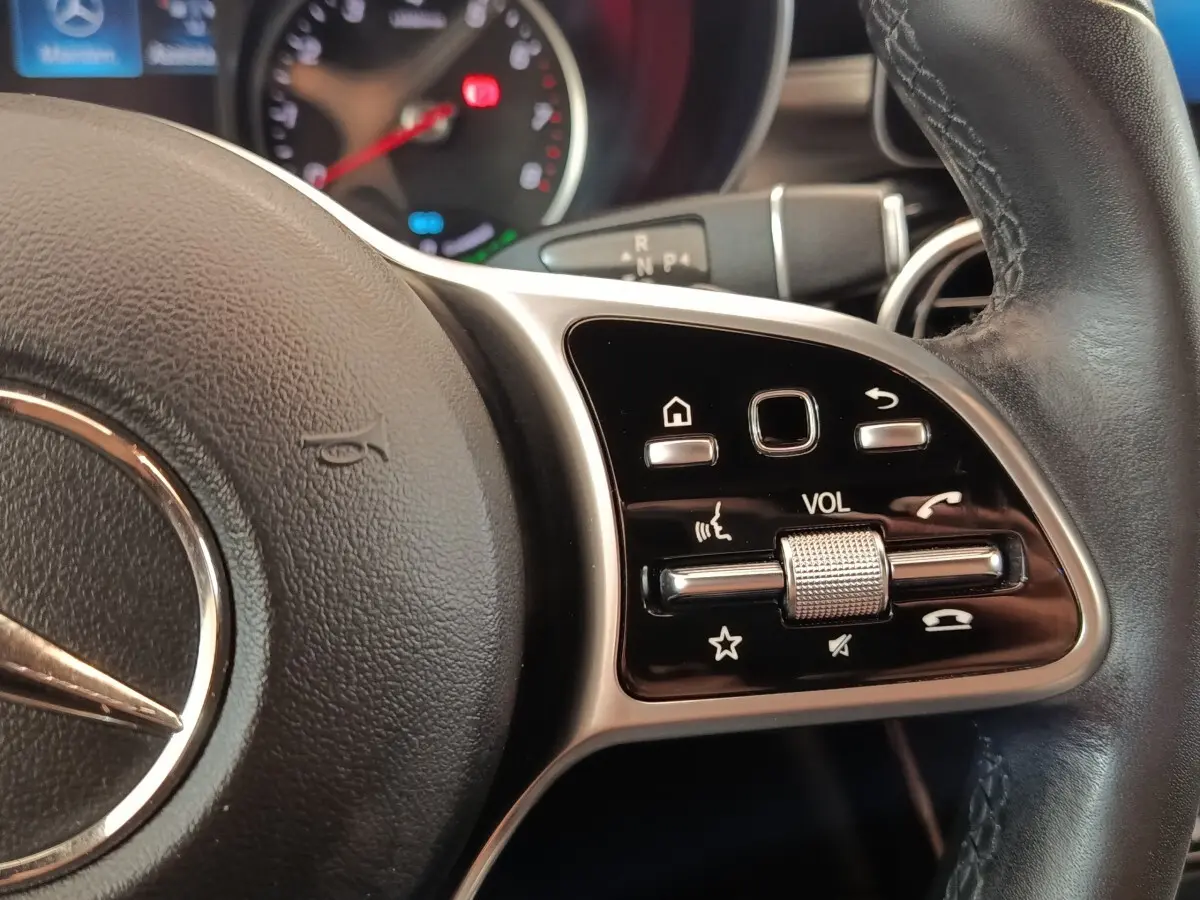 Gros plan sur les commandes de volume et fonctions vocales au volant cuir noir du Mercedes GLC 300 hybride bleu.