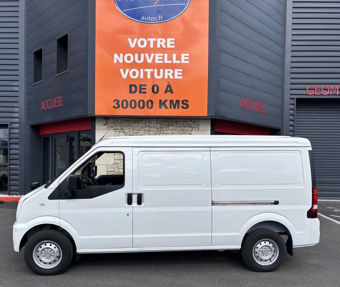 Vue de profil côté gauche d'un utilitaire blanc DFSK EC35 SERES ECOCITY 2023 stationné devant un bâtiment commercial.