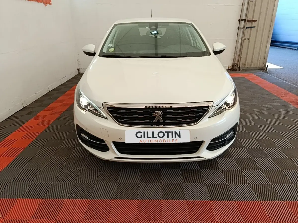 Peugeot 308 blanche vue de face dans un garage, avec calandre chromée et phares allumés.
