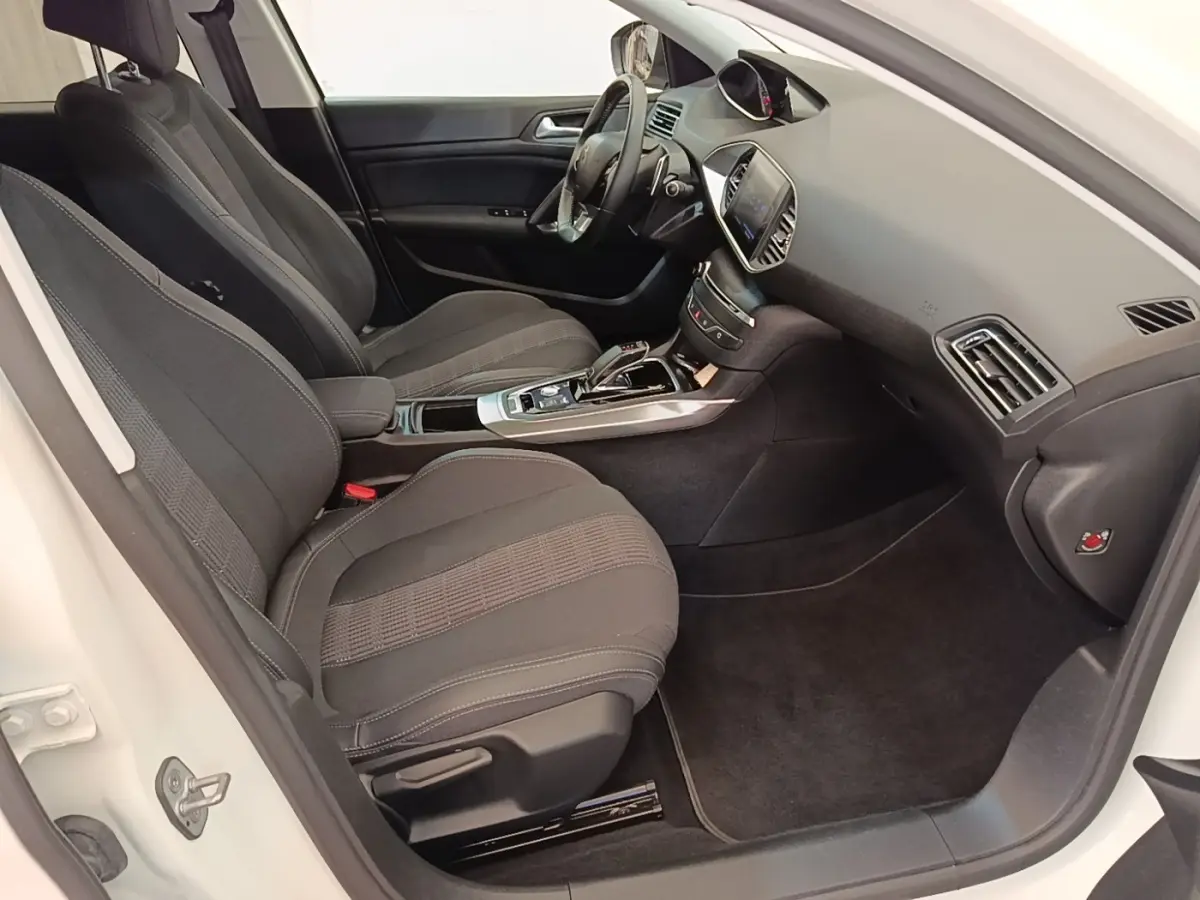Intérieur avant droit de la Peugeot 308 blanche, montrant sièges noirs, tableau de bord avec écran tactile et volant cuir.