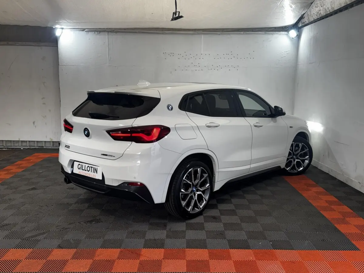 BMW X2 blanc vue 3/4 arrière droit dans un garage, avec jantes alliage et feux arrière LED allumés.