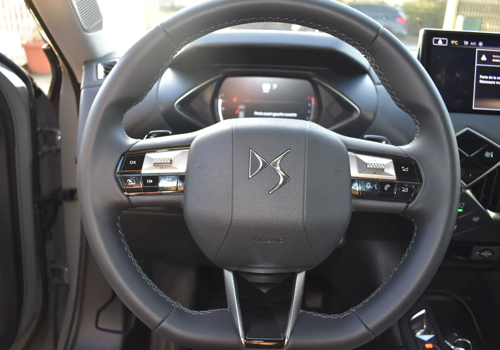 Vue rapprochée du volant noir du DS3 Crossback 2024 avec commandes intégrées et tableau de bord numérique en arrière-plan.