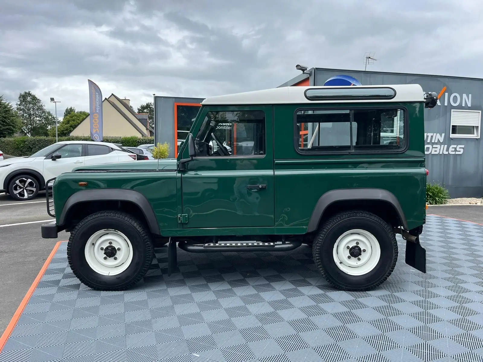 Profil côté gauche d'un Land Rover Defender 90 Hard Top vert avec toit blanc, marchepieds et roues blanches visibles.