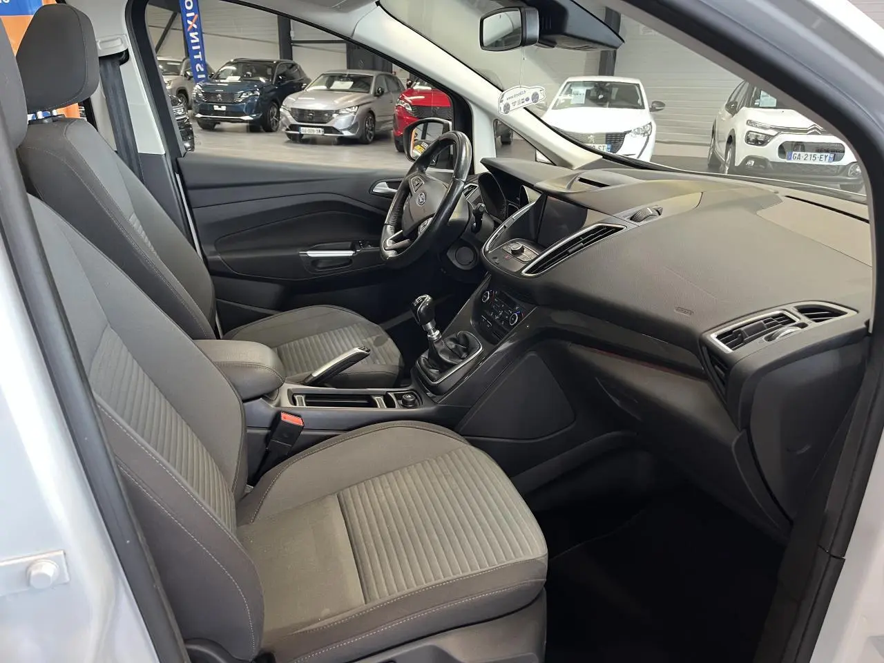 Intérieur avant droit du Ford C-MAX blanc 2018, montrant sièges tissu gris, volant cuir et console centrale avec écran tactile.