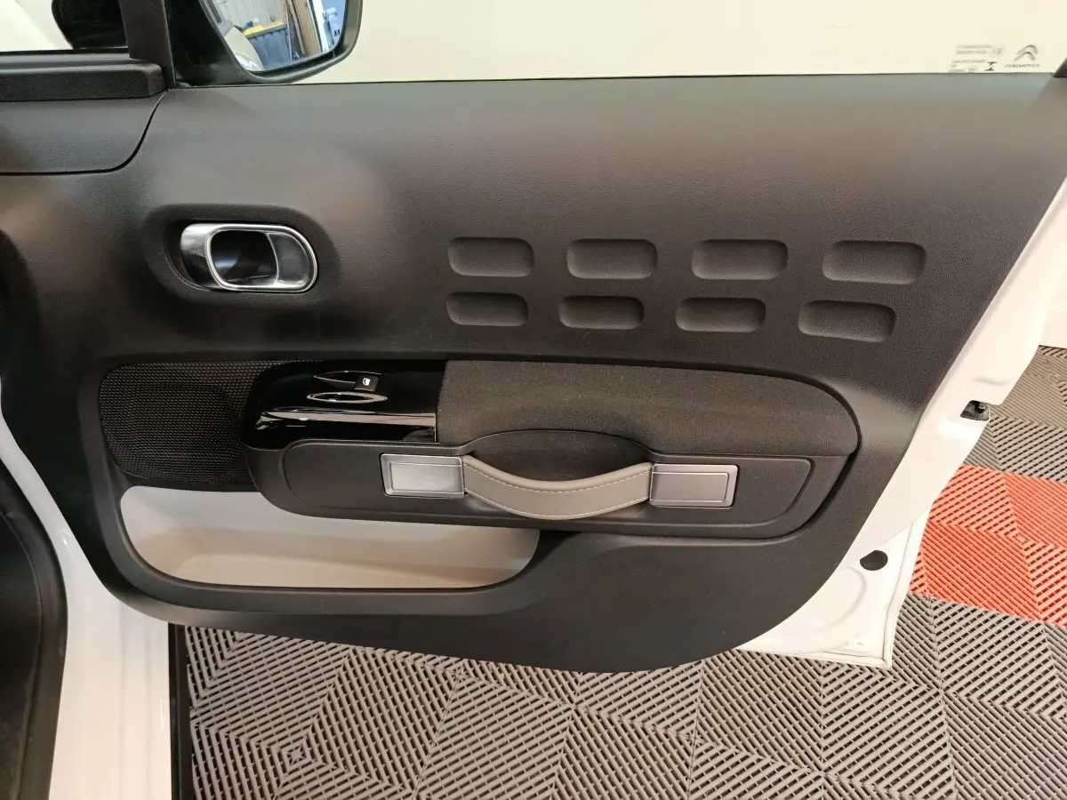 Vue intérieure du panneau de porte avant gauche de la Citroën C3 blanche, avec poignée en cuir et commandes électriques.