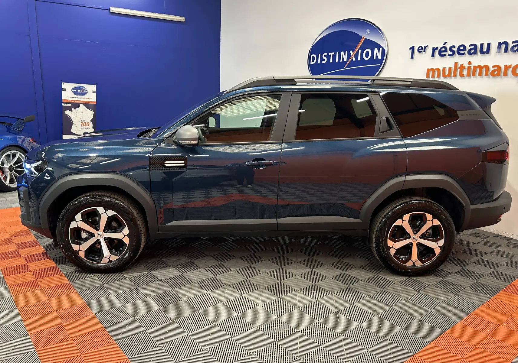 Vue de profil côté gauche du Dacia Bigster bleu indigo avec jantes bi-ton et barres de toit noires en intérieur showroom.