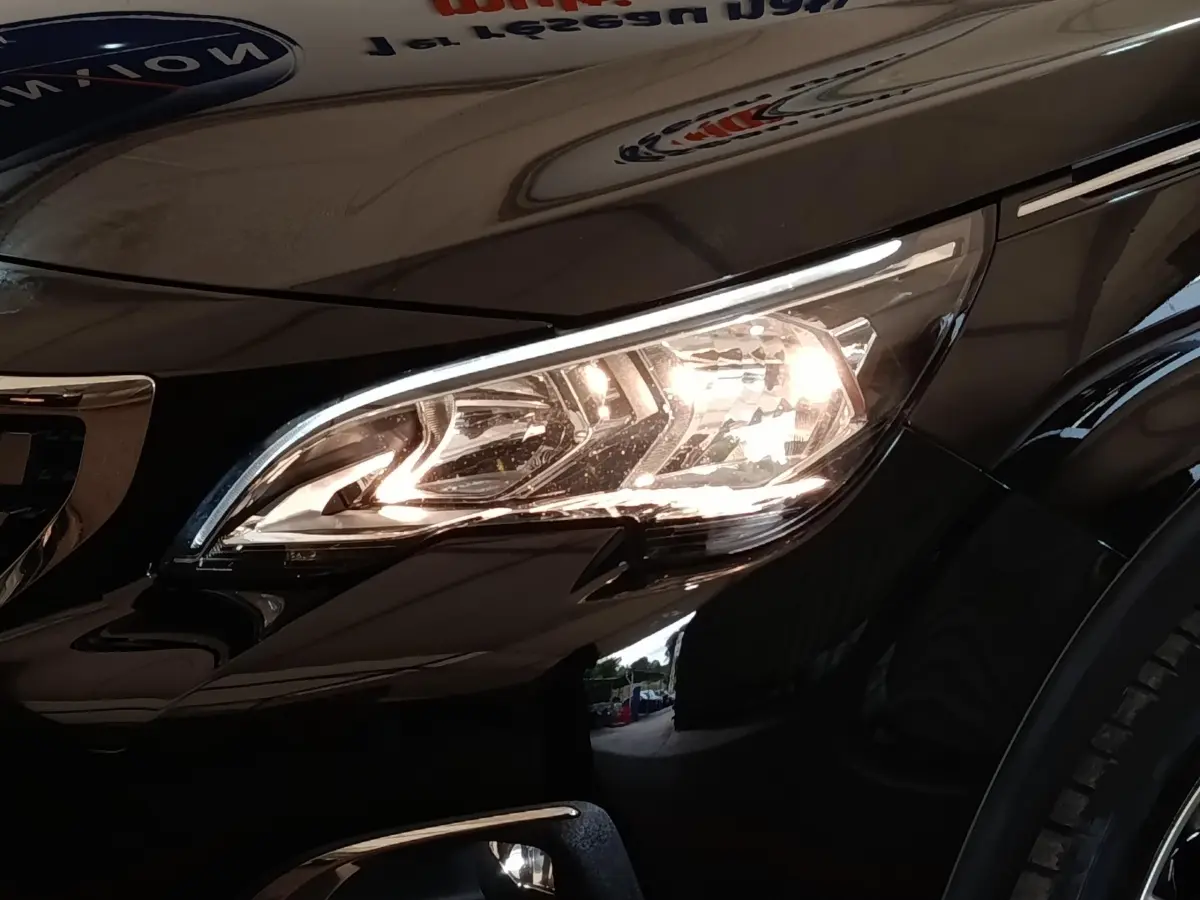 Gros plan sur le phare avant droit allumé d’un Peugeot 5008 noir, avec reflet sur la carrosserie brillante.