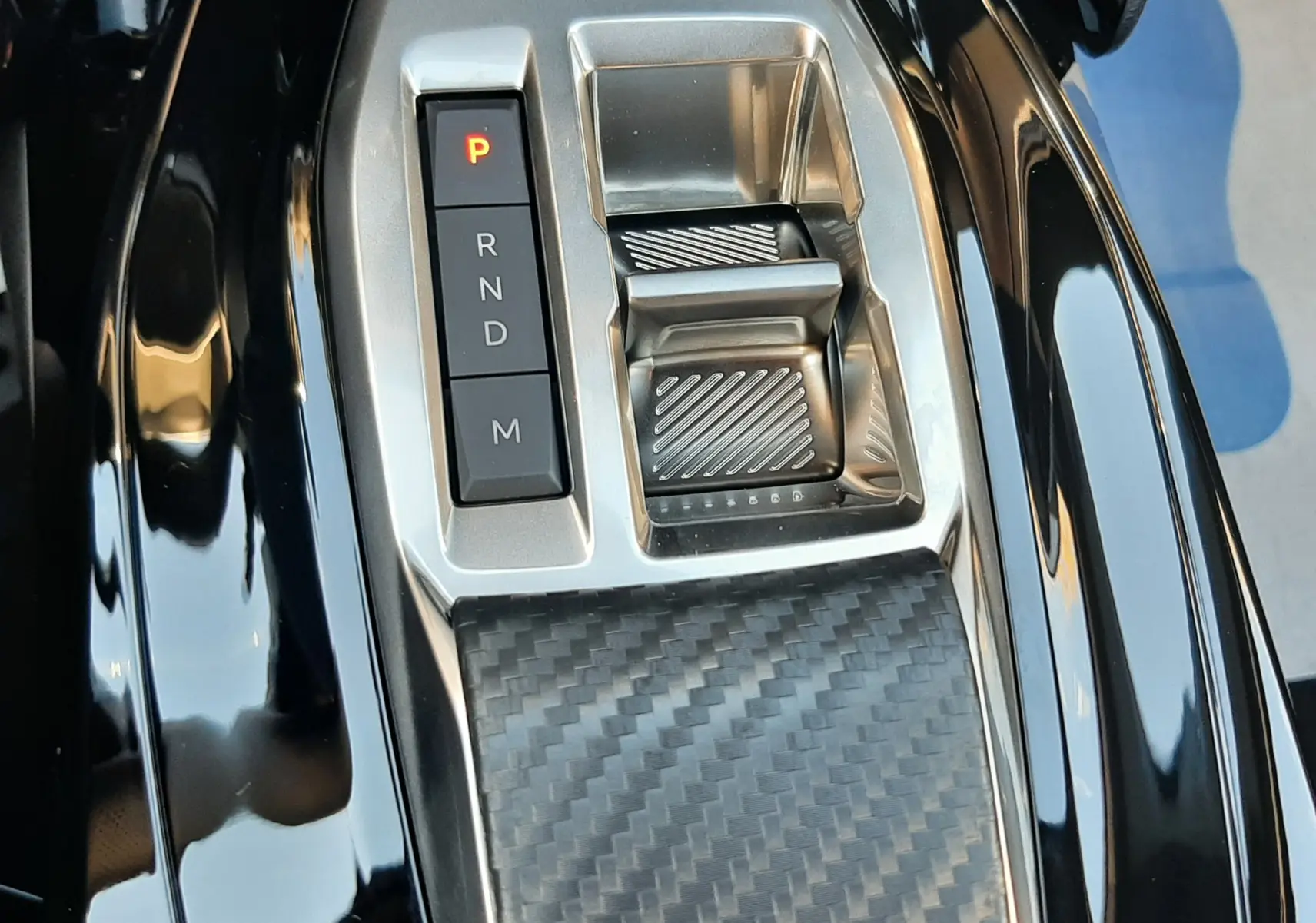 Gros plan sur la console centrale noire brillante du Peugeot 2008 1.2 Hybrid 145ch GT avec sélecteur de boîte auto et finition carbone.