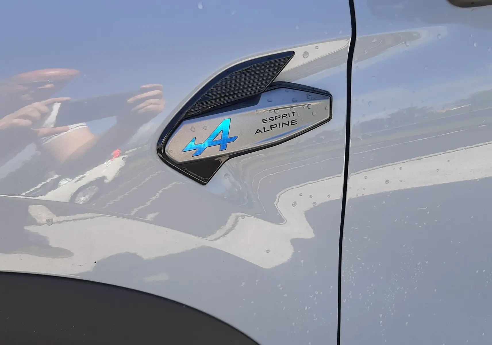 Gros plan sur l’écusson Esprit Alpine bleu sur la porte côté gauche d’une Renault Symbioz gris Rafale.