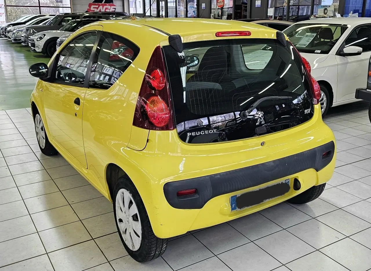 Vue 3/4 arrière droite d'une Peugeot 107 jaune vif en intérieur, avec feux arrière verticaux et pare-chocs noir.