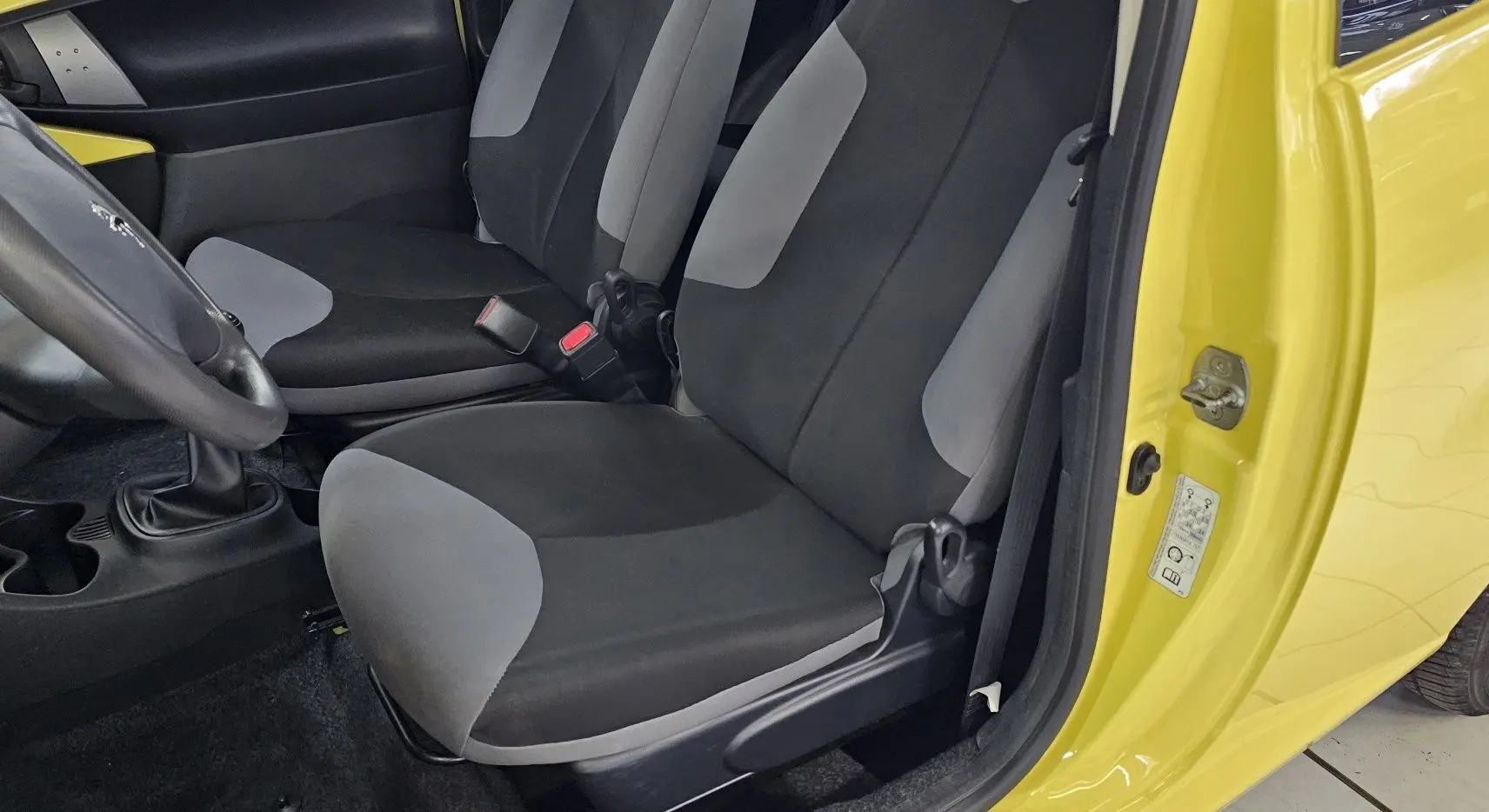 Intérieur côté conducteur d'une Peugeot 107 jaune 2013, sièges tissu noir et gris, volant et levier de vitesses visibles.