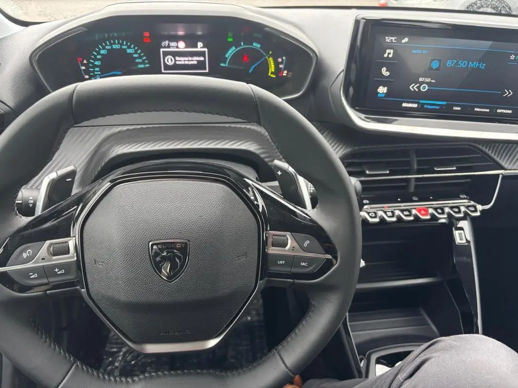 Vue intérieure centrée sur le volant noir de la Peugeot 208 Hybrid 100 e-DCS6 Style avec tableau de bord numérique et écran tactile.