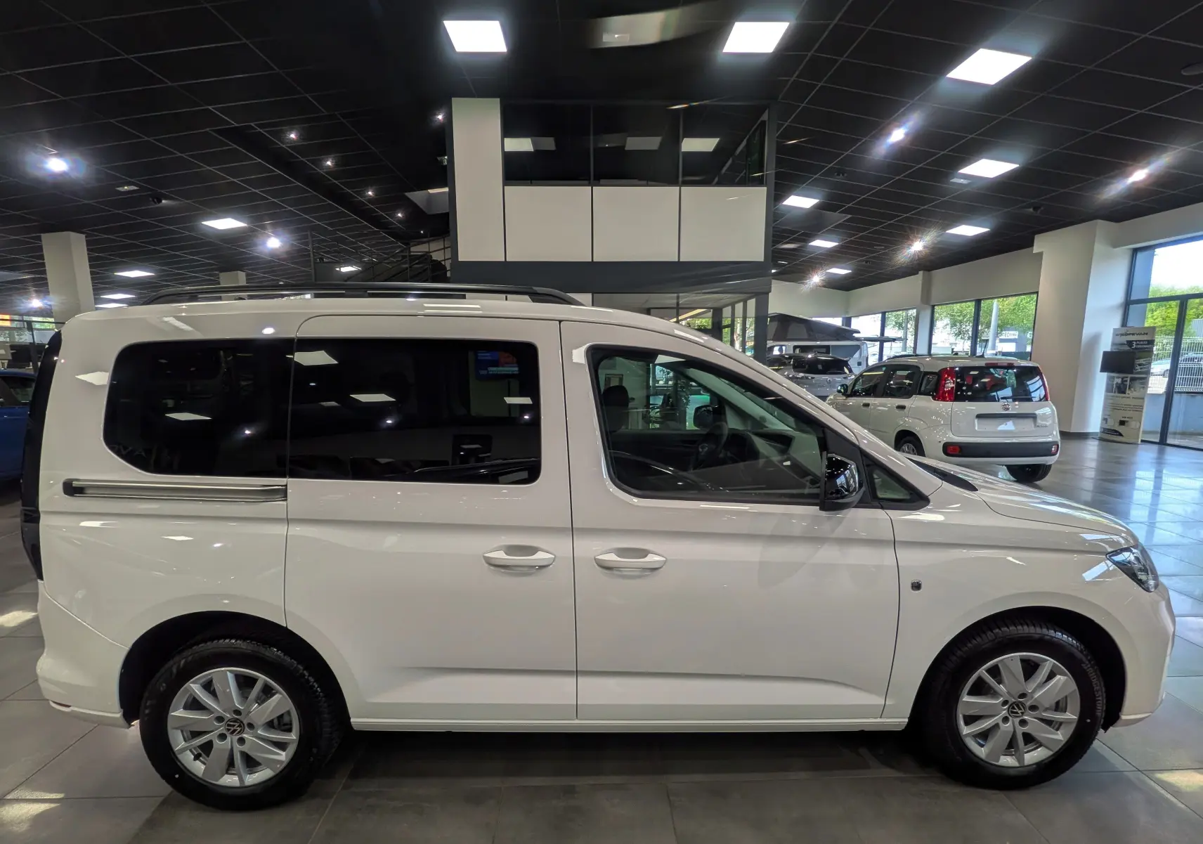 Vue de profil côté droit d'un Volkswagen Caddy blanc 2025 en showroom, avec vitres arrière teintées et jantes alliage.