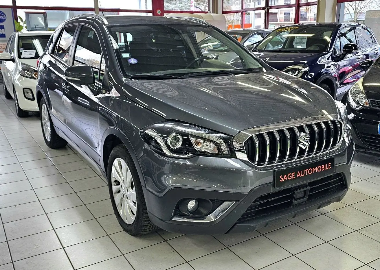 Suzuki SX4 S-Cross gris en 3/4 avant droit, avec calandre chromée et antibrouillards intégrés, en showroom.