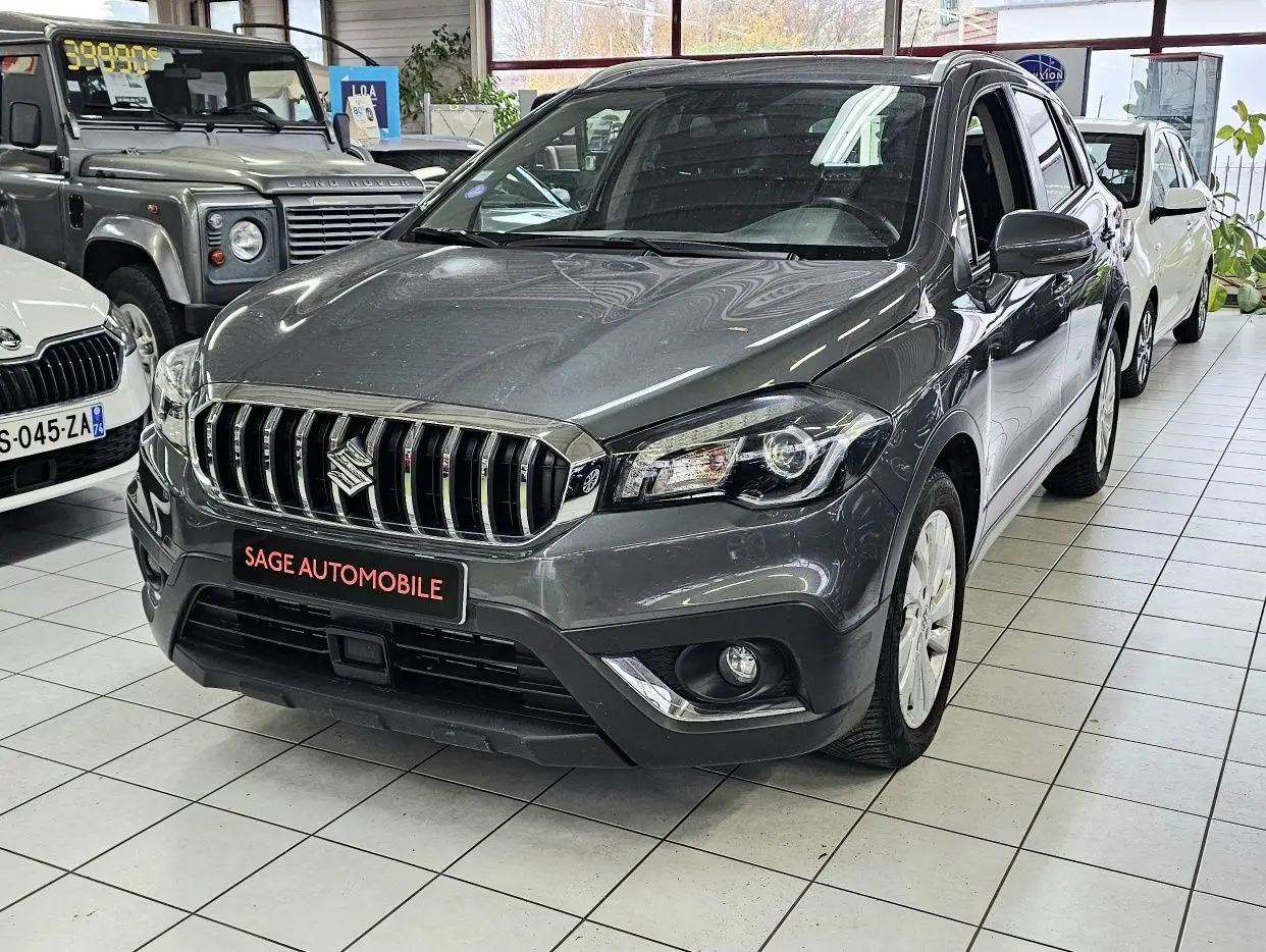 Suzuki SX4 S-Cross gris vu de face droite en showroom, avec calandre chromée et phares LED visibles.