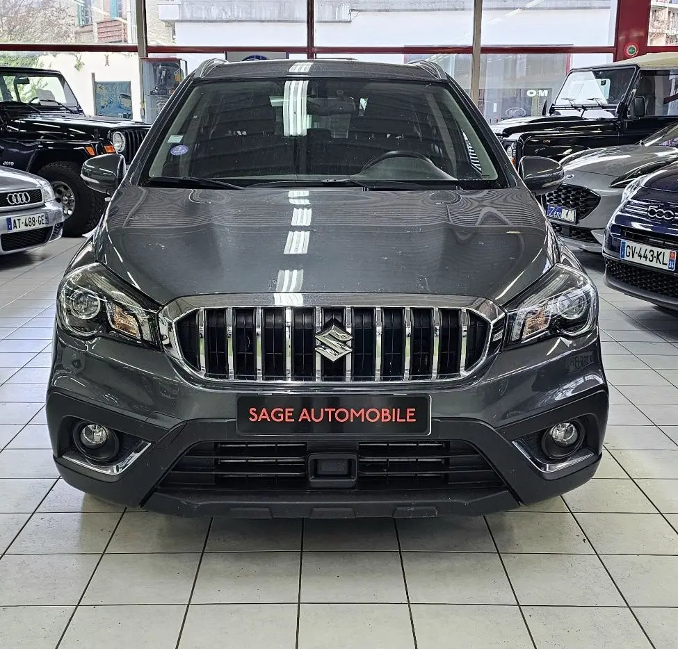 Vue frontale d'un Suzuki SX4 S-Cross gris exposé en showroom avec calandre chromée et phares allumés.