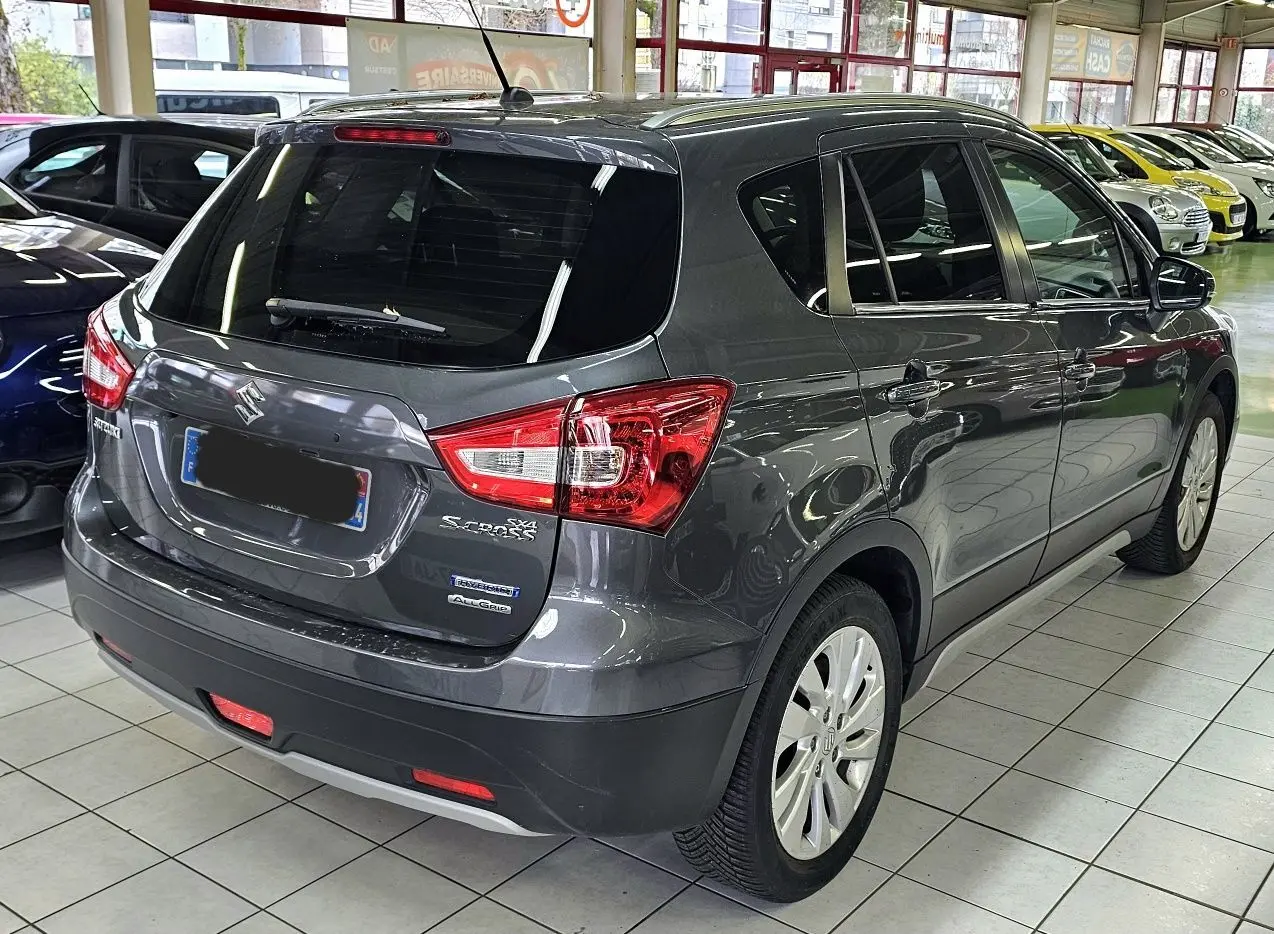 Vue 3/4 arrière droite d'un Suzuki SX4 S-Cross gris métallisé en showroom, avec badges Hybrid et AllGrip visibles.