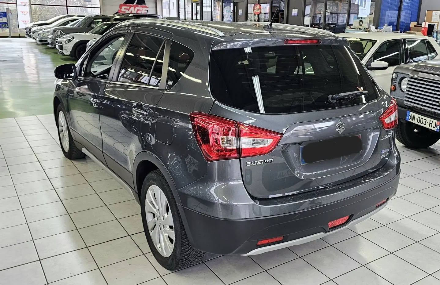 Suzuki SX4 S-Cross gris en 3/4 arrière droit, stationné en intérieur avec plusieurs autres véhicules visibles.