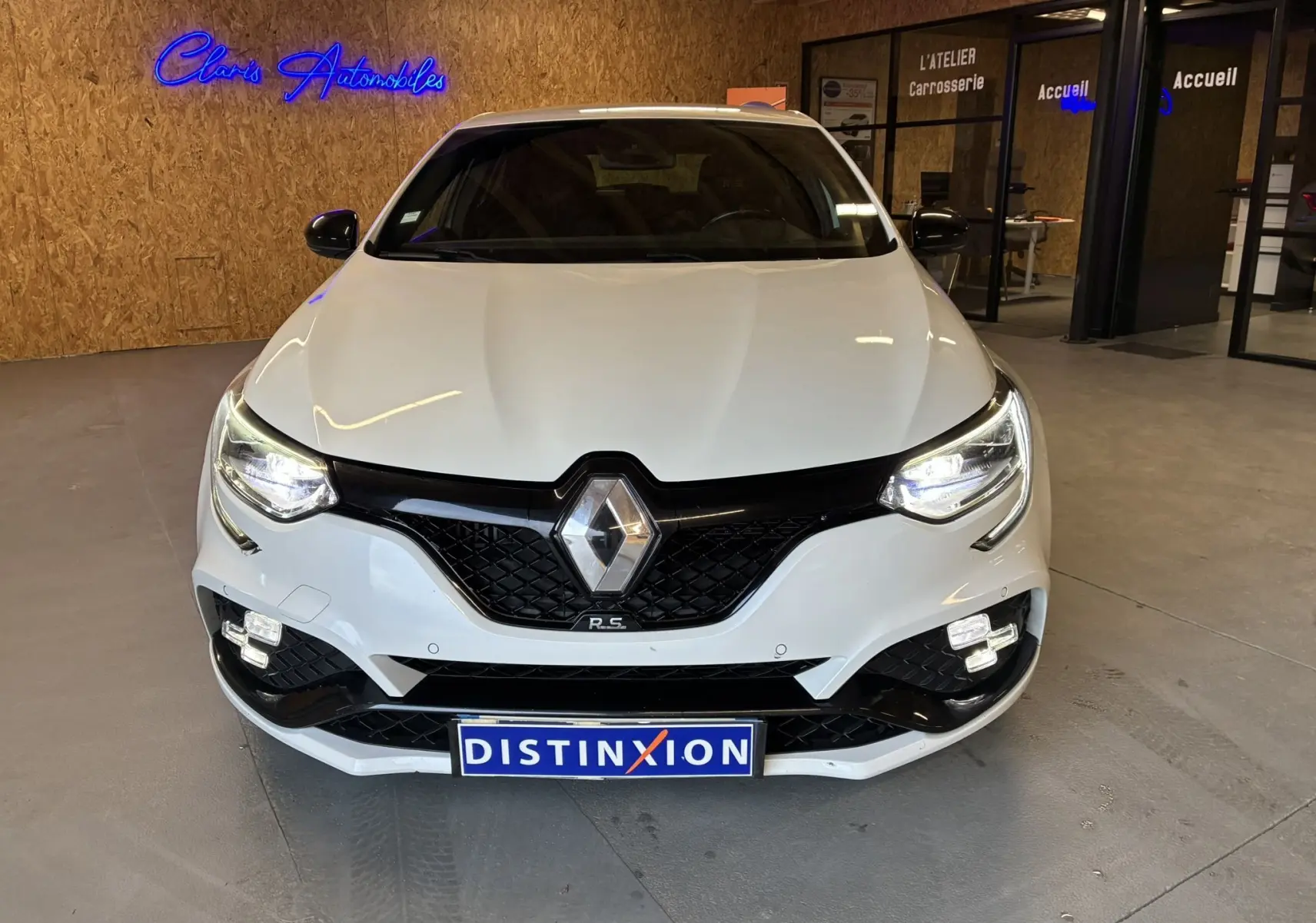 Vue avant d'une Renault Mégane IV RS 280 EDC6 blanche avec calandre noire et phares allumés en intérieur showroom.
