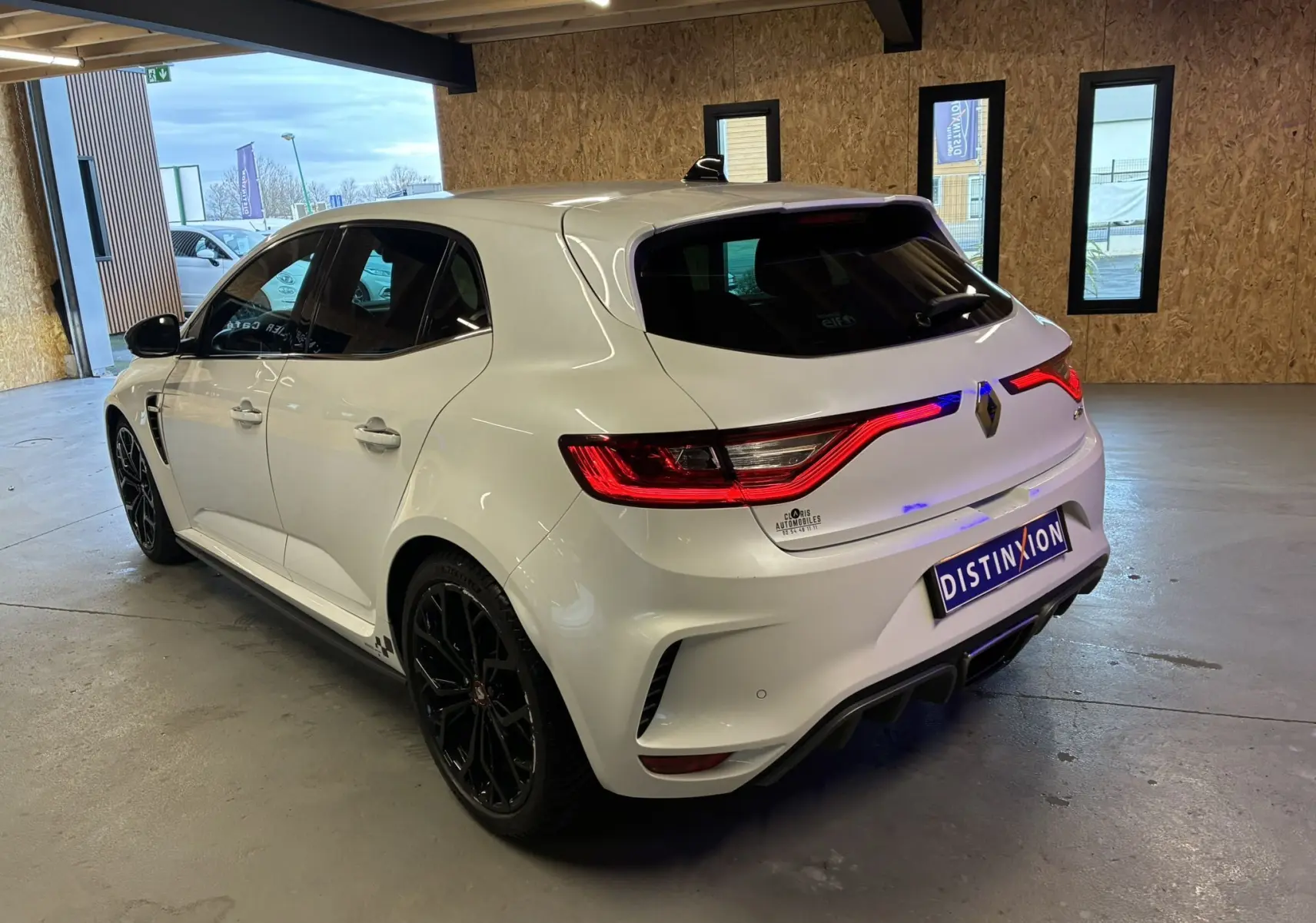 Vue 3/4 arrière droite d'une Renault Mégane IV RS 280 EDC6 blanche avec jantes noires et feux arrière allumés en intérieur.