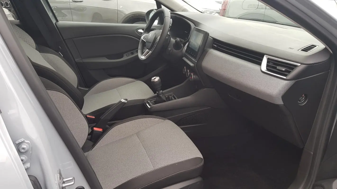 Vue intérieure côté conducteur de la Renault Clio gris 2025, montrant sièges tissu, tableau de bord et levier de vitesse manuel.